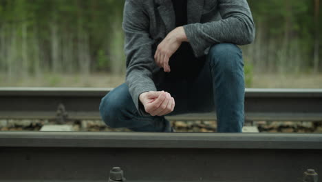 a close-up view of a man in a grey blazer and blue jeans kneeling by a railway track, carefully touching the rail with his hand, then removed the hand from the trail track