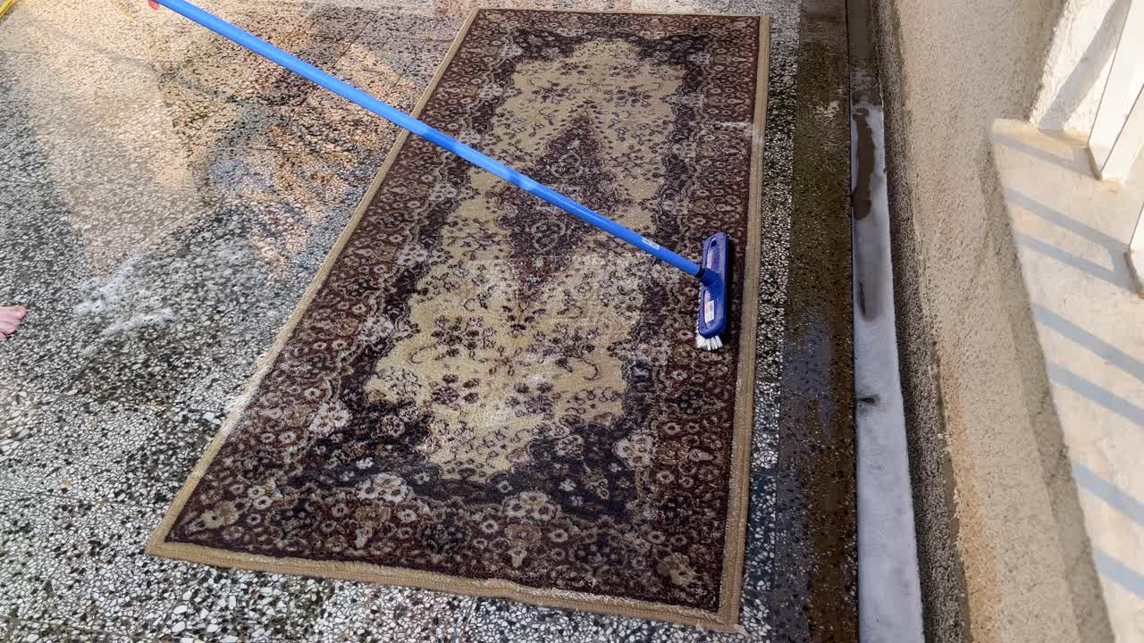 A woman washes brown carpet by hand, manually on the terrace with a long stick brush