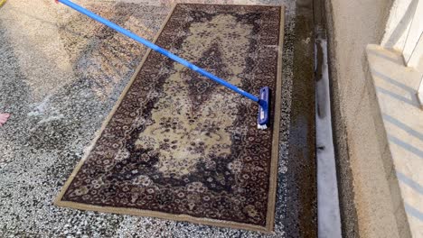 una mujer lava la alfombra marrón a mano, manualmente en la terraza con un cepillo de palo largo