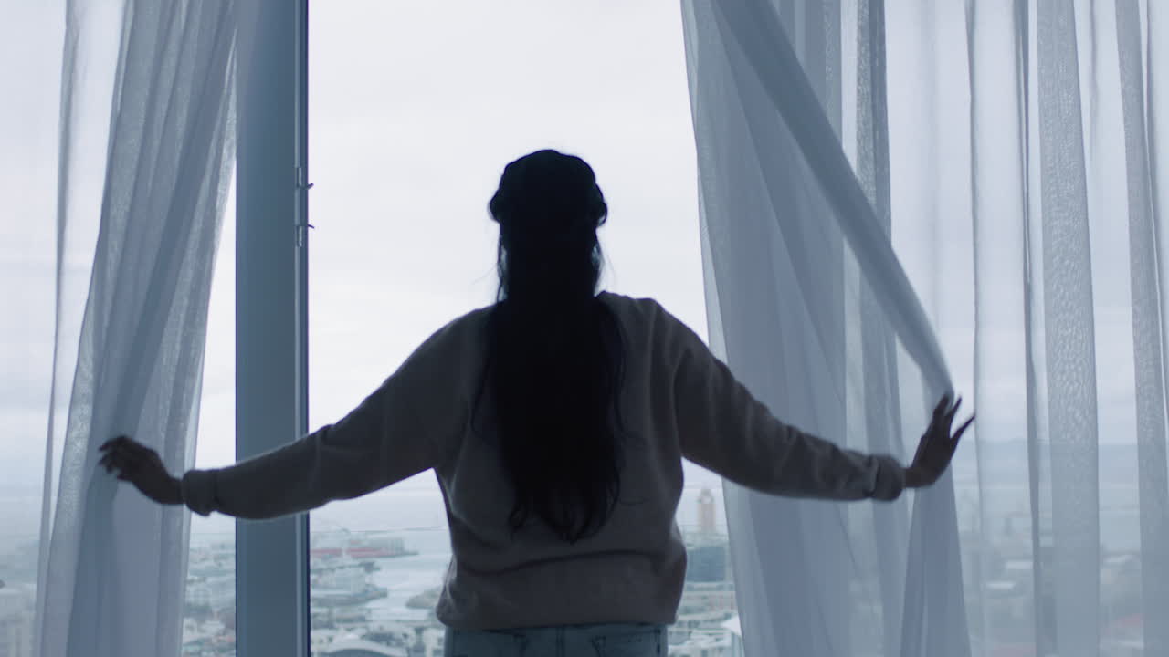 행복한 젊은 여성은 커튼을 열고 창문 밖을 바라보며 신선한 새로운 날을 준비하고 휴식을 취하고 도시의 전망을 즐기고 있습니다.
