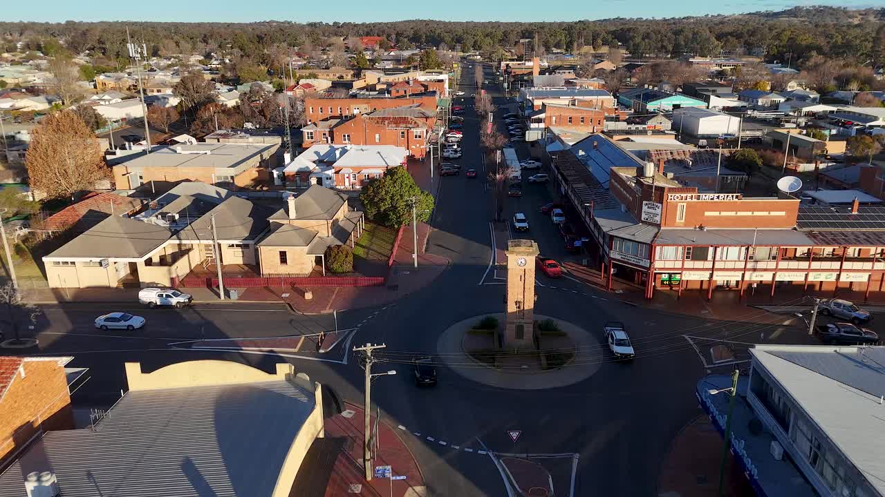 Aerial drone footage glides above a small town’s main street, circling a central clock tower roundabout with late afternoon sunlight and light traffic