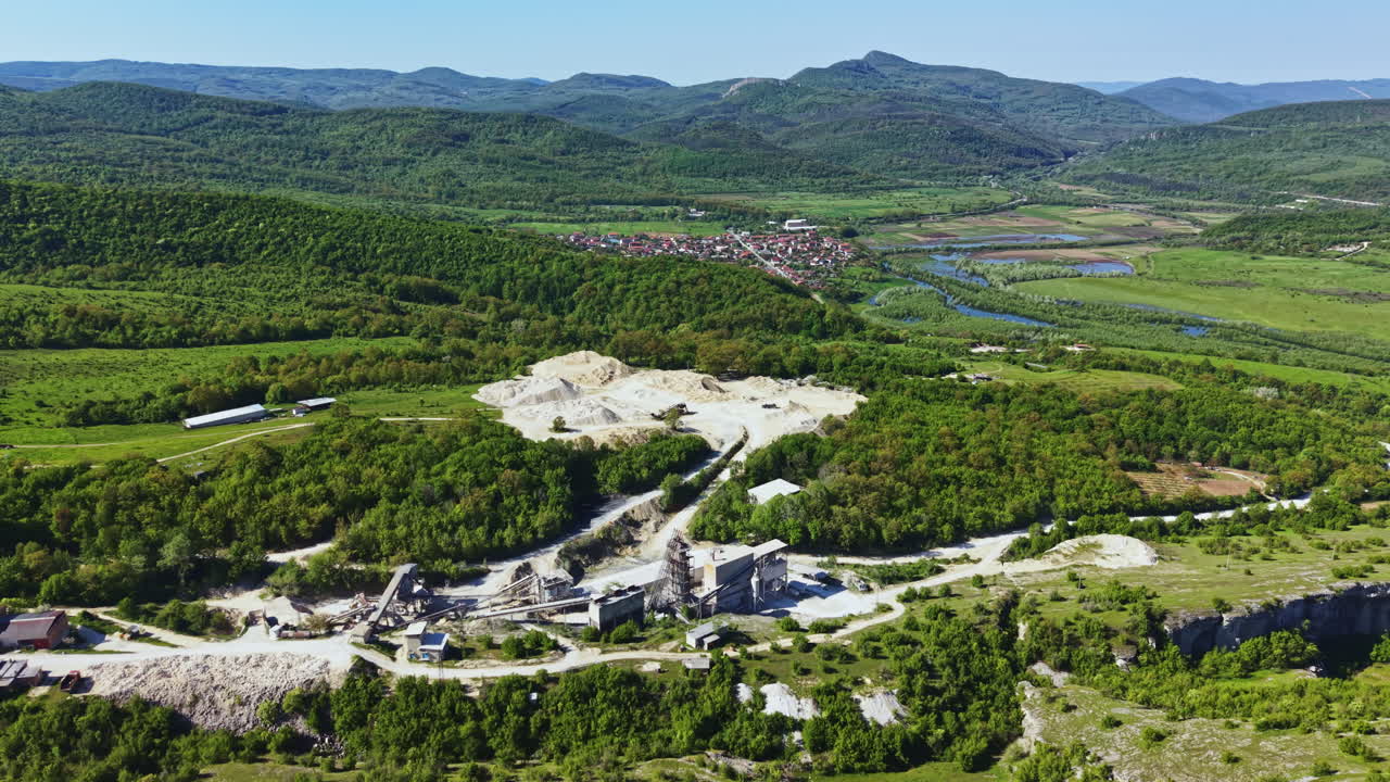 Ett stort stenbrott ligger i ett grönt landskap med skogsområden och berg i bakgrunden, beläget nära en liten stad en klar dag.
