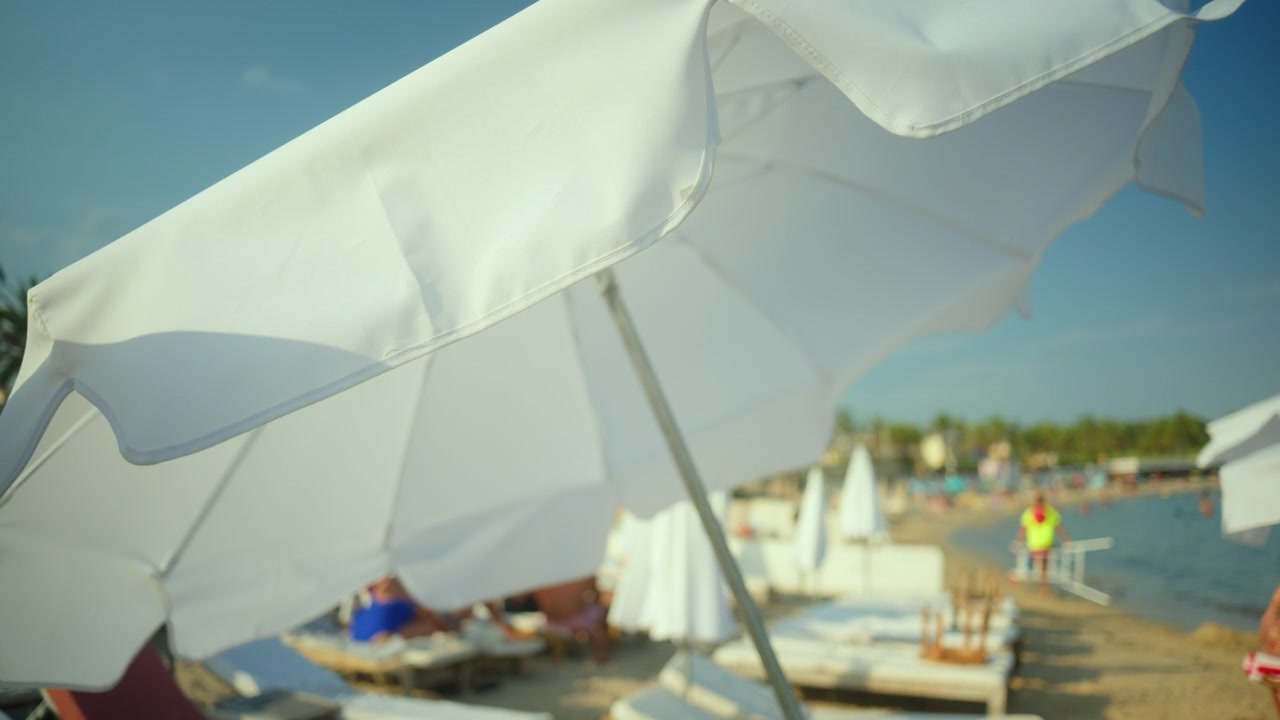 Ombrellone bianco da spiaggia che ripara dal sole in un affollato stabilimento balneare. Persone che si rilassano su sedie a sdraio e nuotano nel mare blu.