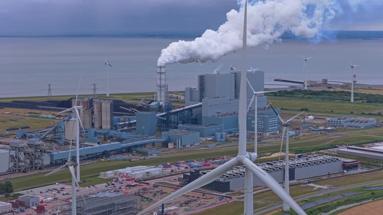 Aerial view of a modern industrial complex with spinning wind turbines contrasting with a power plant releasing a plume of smoke
