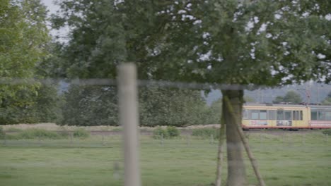 A-single-car-yellow-tram-with-a-black-roof,-moving-from-left-to-right-in-a-rural-setting,-numbered-"5"-on-its-destination-sign,-set-against-a-backdrop-of-a-grassy-field,-trees,-and-a-distant-hill