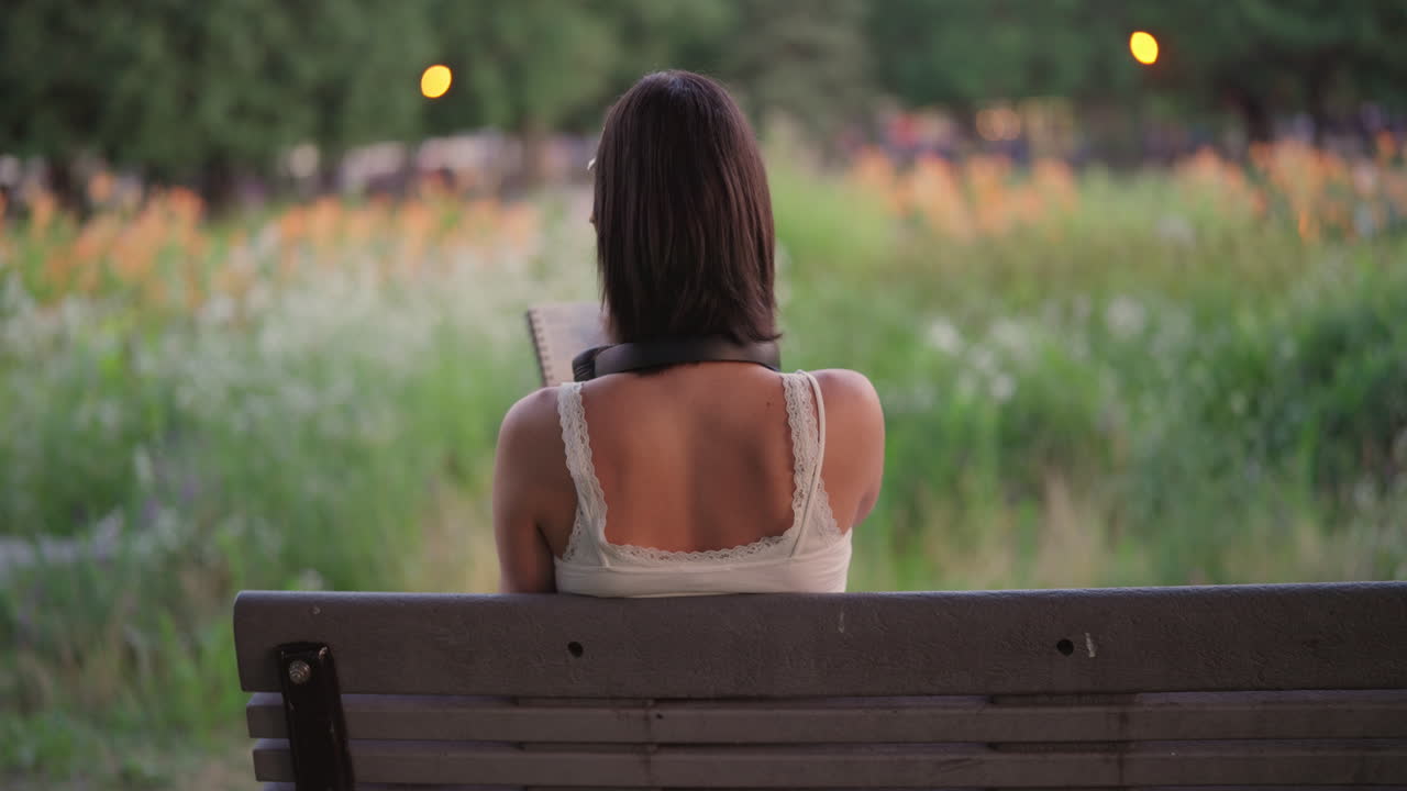 Woman with headphones sits on bench, journals and writes in notebook alone, slow pan left