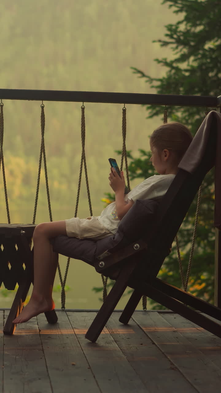 niños pequeños juegan juegos en teléfonos inteligentes en la terraza transparente al atardecer. hermano y hermana descansan en cómodos sillones disfrutando de una increíble vista del denso bosque
