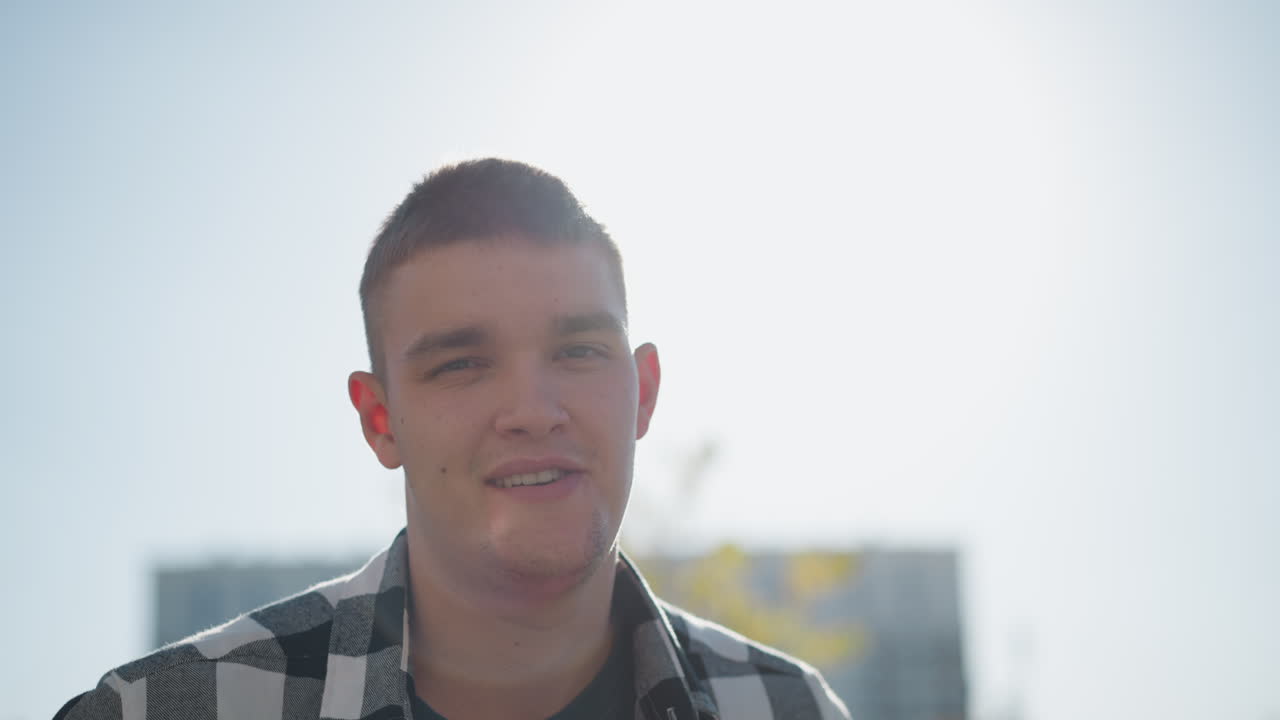 handsome man adjusts checkered shirt while smiling warmly under bright sky with sunlight creating radiant halo effect around head against blurred urban background in casual outdoor setting
