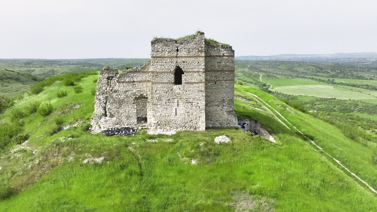 Drone pull-back reveals a lone hilltop castle above lush spring meadows and rolling fields under an overcast sky in rural Bulgaria