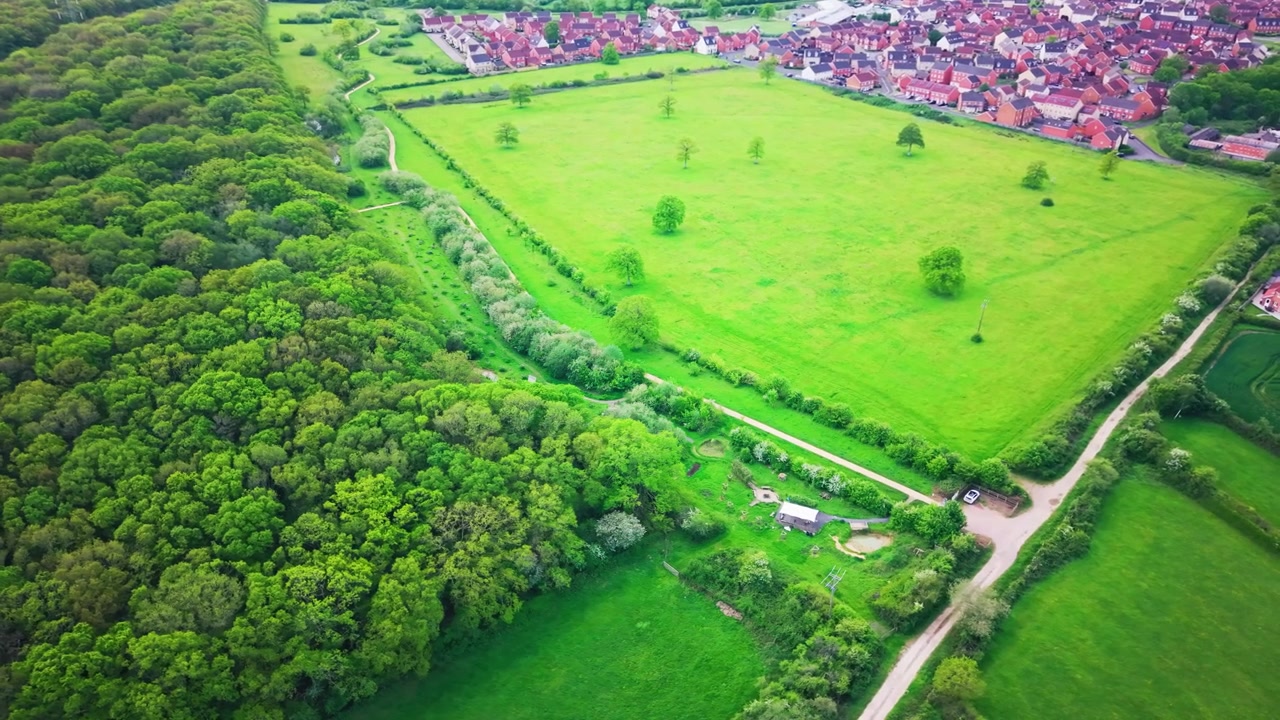 Letecké záběry dronem ukazující okraj lesa, zelený koridor, otevřené travnaté plochy, pěšiny, živé ploty, stromy a nedaleké sídliště na okraji města v Anglii.