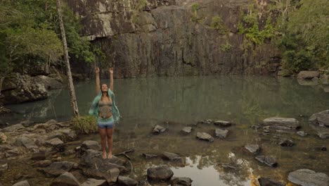 Chica-Con-Bufanda-De-Pie-Sobre-Una-Roca-Y-Levantando-Las-Manos-En-El-Aire---Cedar-Creek-Falls-En-Qld,-Australia