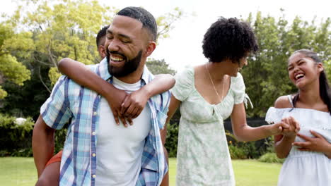 Happy-african-american-parents,-daughter-and-son-piggybacking-and-walking-in-garden,-slow-motion