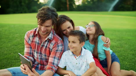 family sitting on grass in park. parents and children making video call with pad