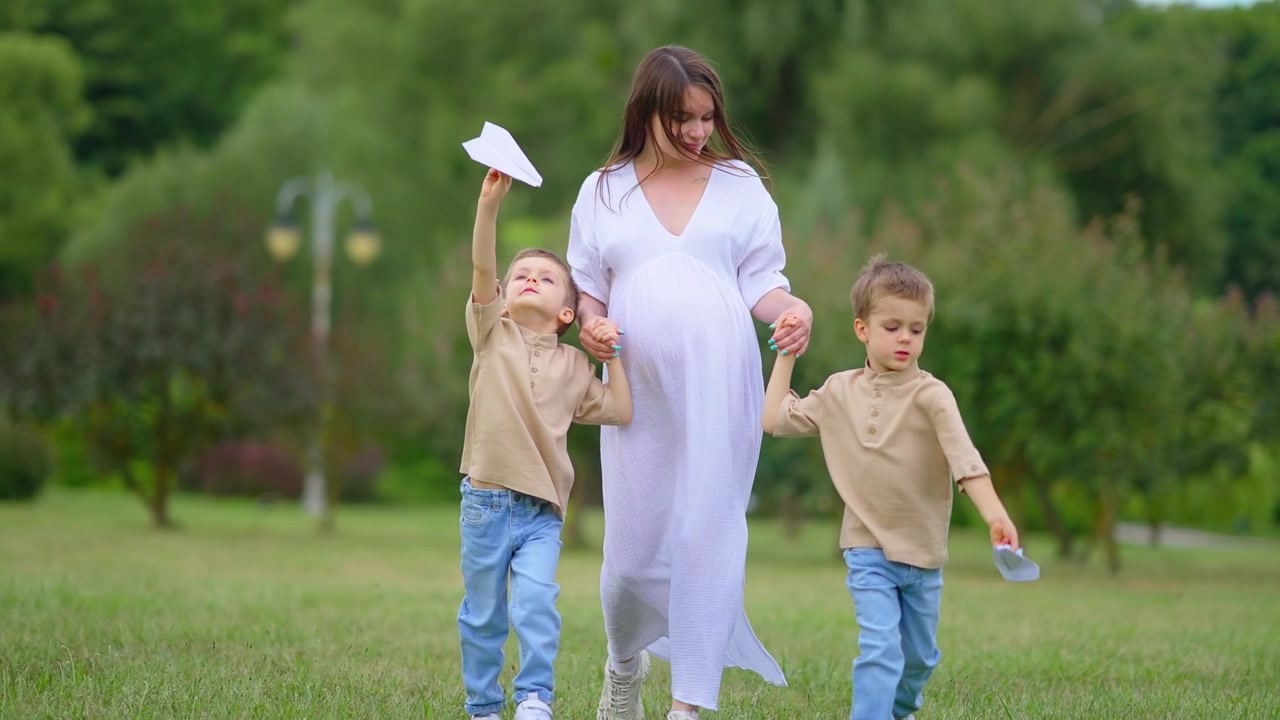 Zwangere vrouw loopt met twee jonge jongens die papieren vliegtuigjes vasthouden in het park