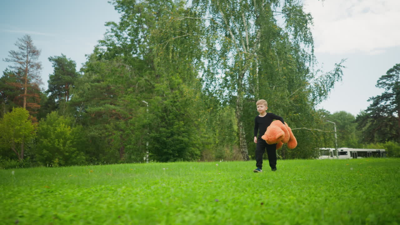 Fair skin boy dressed in black walking across vibrant green park holding large plush teddy bear, surrounded by trees and distant parked buses under soft daylight in peaceful natural outdoor setting