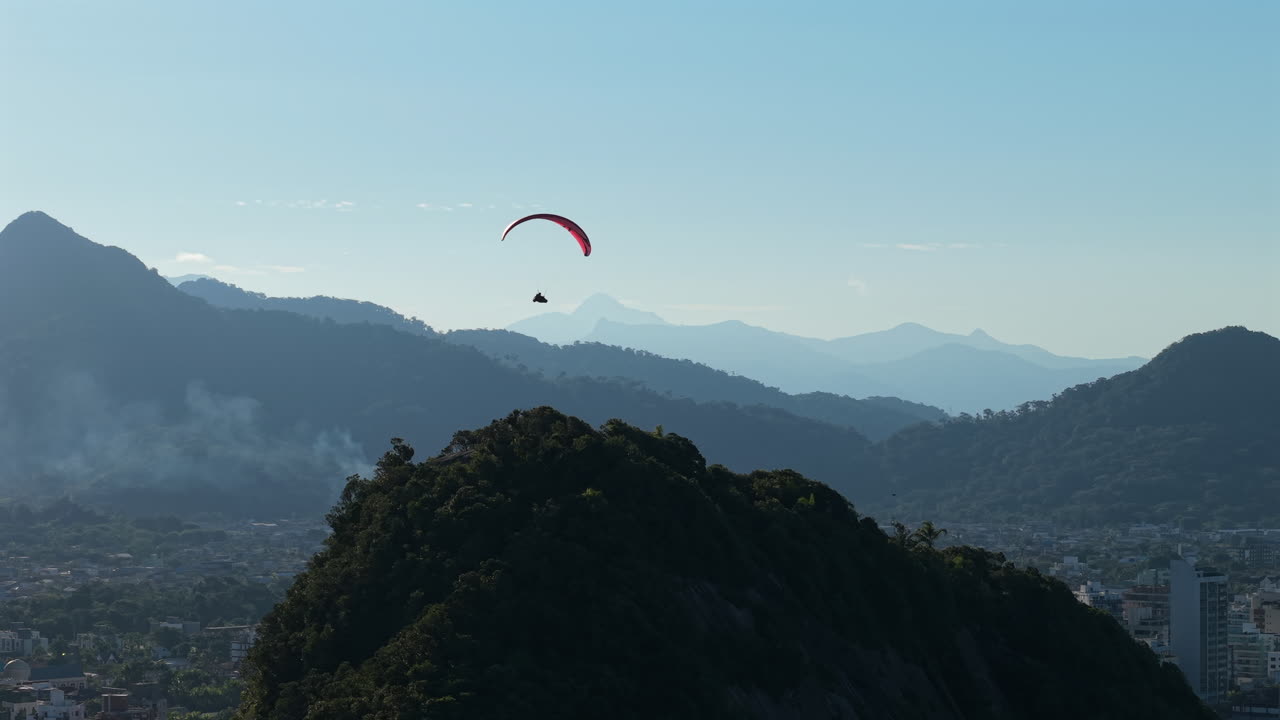 A stunning aerial parallax tracking shot follows a paraglider soaring peacefully over the beautiful mountains and city of Caiobá, Brazil, on a clear day