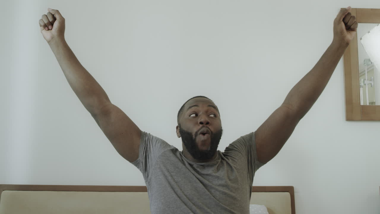 Young guy outstretched arms in bed in morning. Black man dancing in bed.