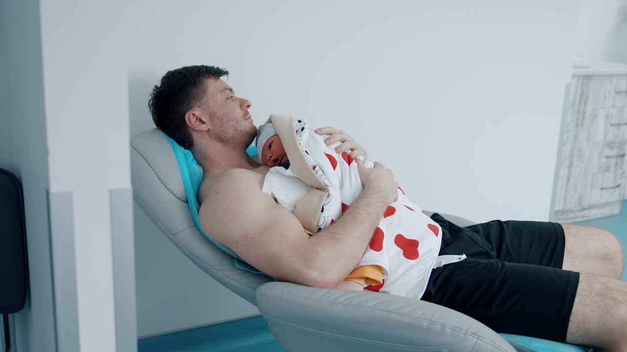 Caring father is holding newborn baby. Dad soothes his child after birth while mother is resting after C-section.