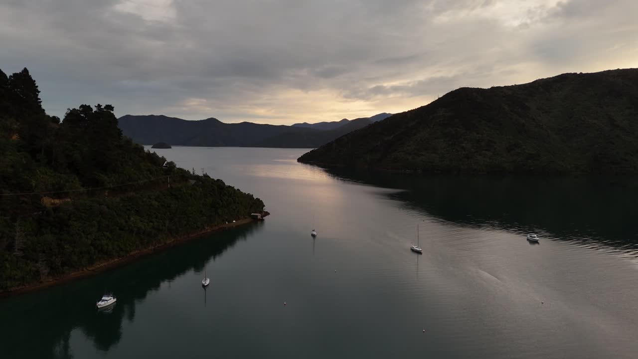 Several sailing boats and speedboats on lake between mountains during. Cloudy sunset. Aerial flyover shot. Picton area in New Zealand. Wide shot
