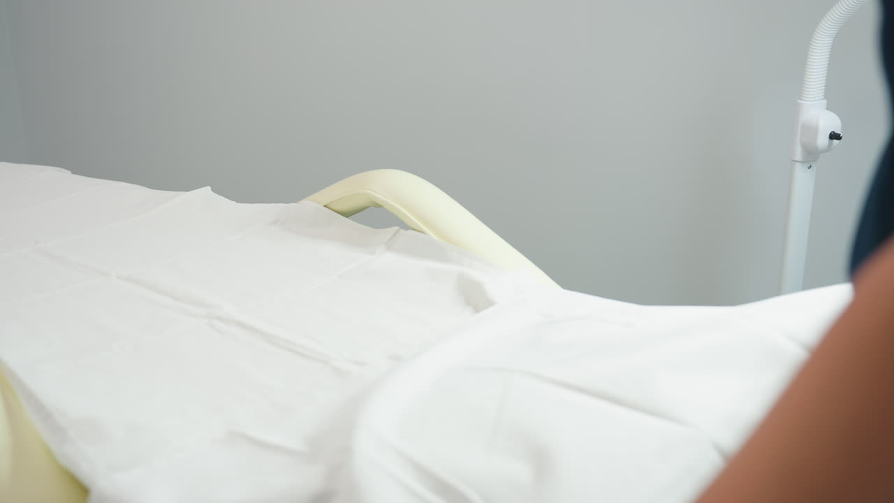 Skin care expert in navy uniform sprays antiseptic onto cotton pad and carefully wipes down pale massage chair in pristine white treatment room, ensuring hygienic surface and welcoming environment