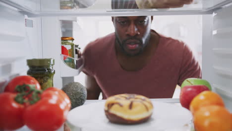 view looking out from inside of refrigerator as man opens door and reaches for unhealthy donut