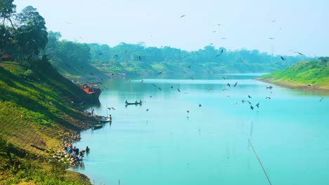 Static-shot-of-river-with-eagles,-hawks-flying-on-Surma-river-with-a-fishing-boat-in-Sylhet,-Bangladesh