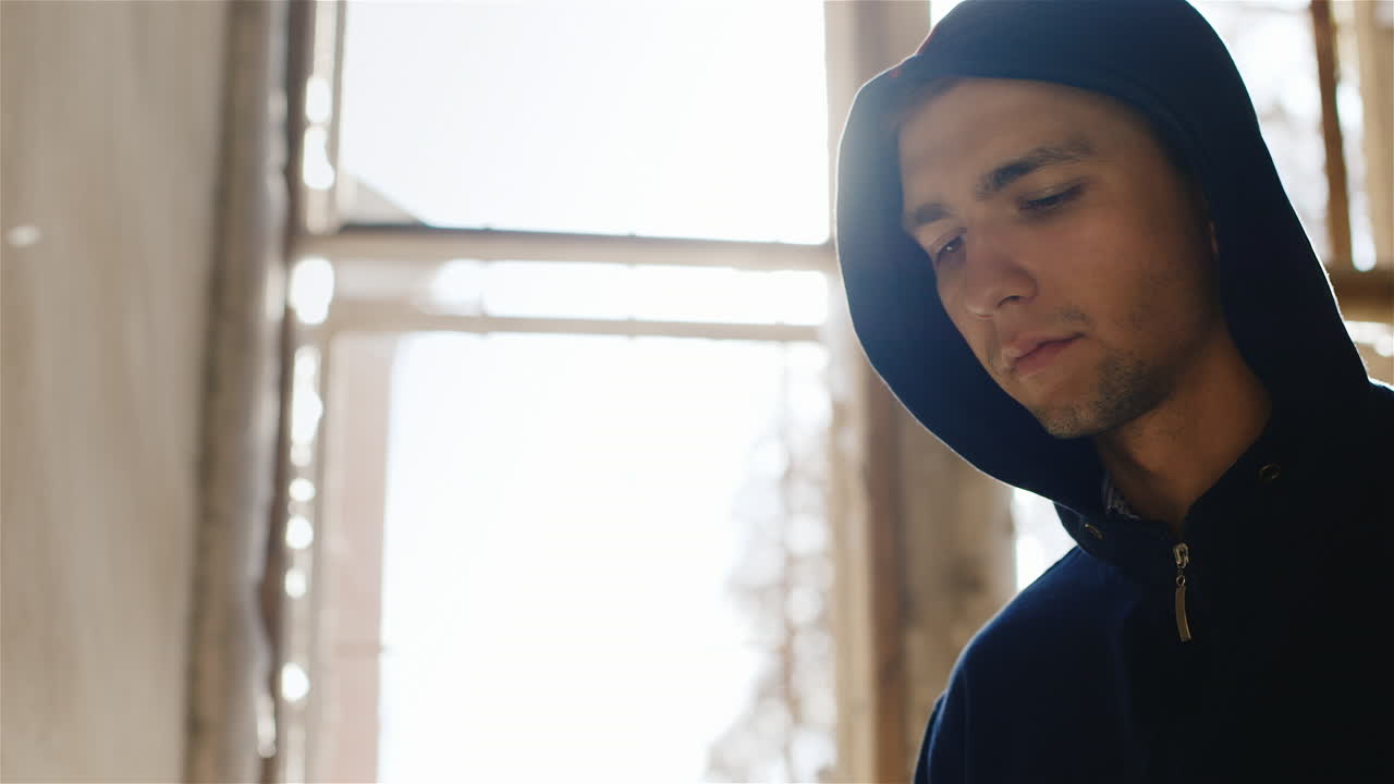 un joven encapuchado fumando un cigarrillo en una casa abandonada cerca de la ventana