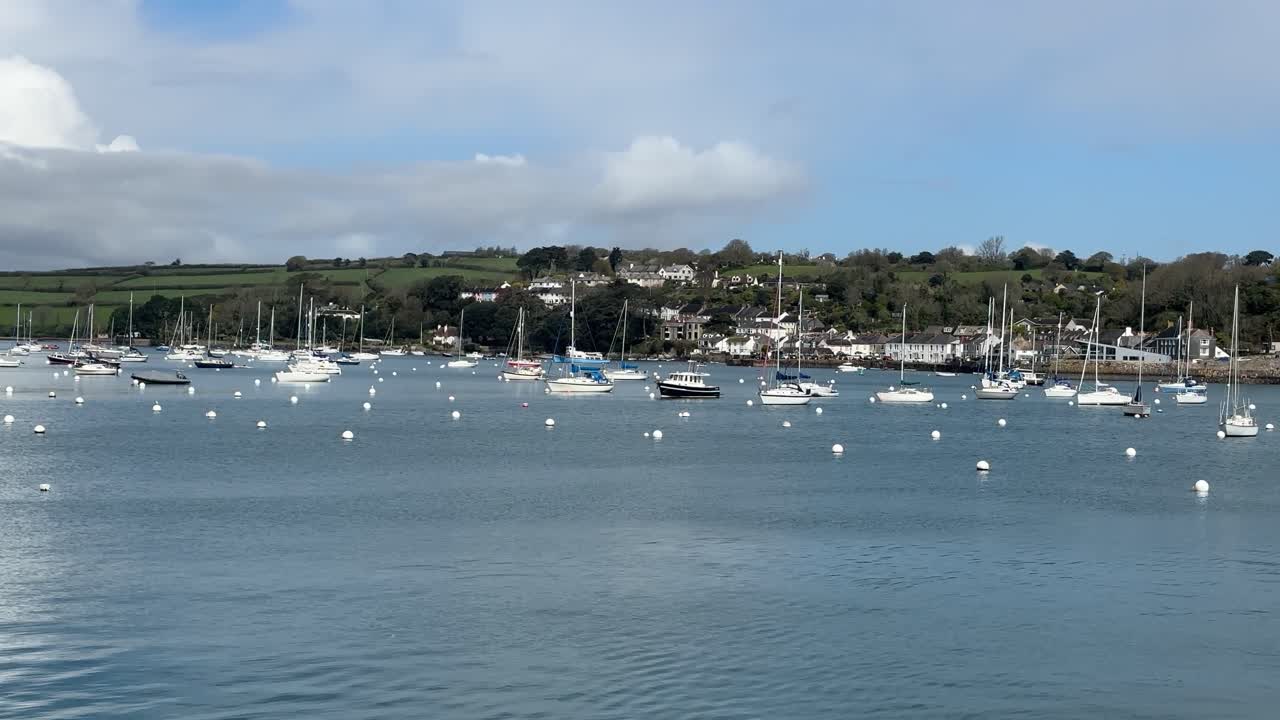 Ampla tomada panorâmica do porto de Falmouth Water na Cornualha, Inglaterra, com muitos veleiros ancorados em águas calmas, colinas verdes ondulantes e casas agrupadas ao fundo sob céus de luz suave do dia.