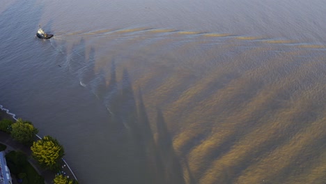 Aerial-birds-eye-view-tugboat-passing-by-a-commercial-river-causing-waves-on-a-twilight-sunset-muddy-water-just-returning-to-the-port-dock-and-passing-a-luxury-lush-residential-boardwalk-community