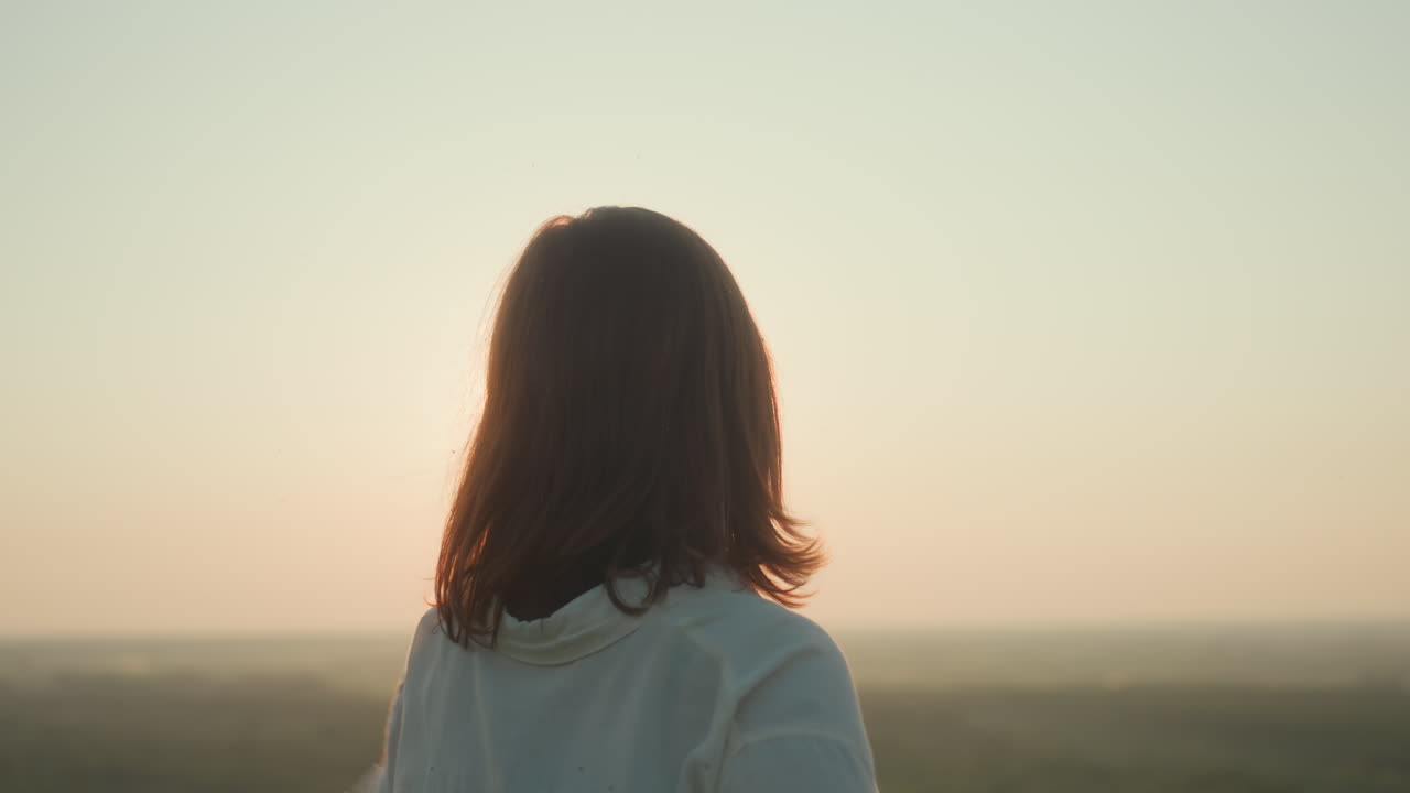 Cheerful lady with closed eyes and gentle smile opens arms under warm sunset glow, sunlight creating lens flare across white shirt as she embraces peaceful outdoor moment