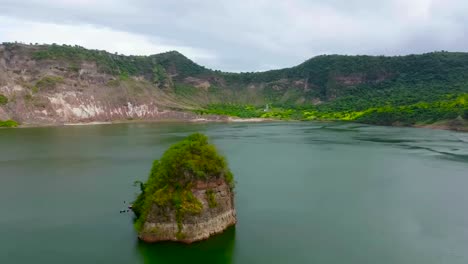 Aerial drone footage gliding over Taal lake or Bombon lake water and focusing on a large rock formation or a small island in the water during a cloudy day in the Philippines. Large mountains visible.