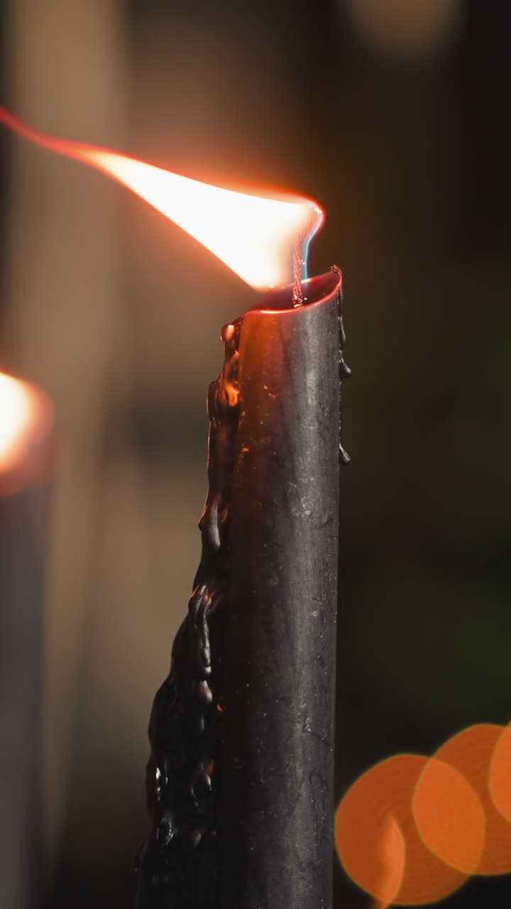Flame of black candle waves in wind on blurry background closeup. Burning symbol of grief and loss in semi-dark room. Funeral ceremony accessory