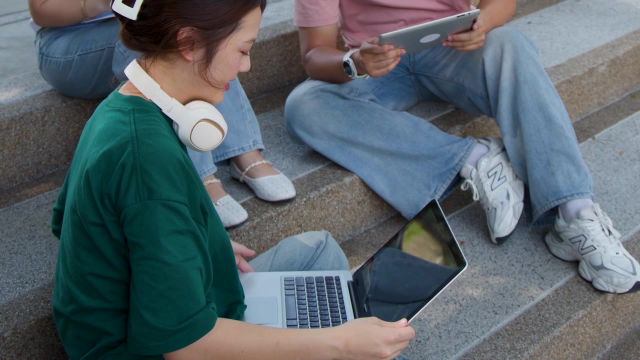 Diverse college students study together outdoors using digital devices on concrete campus stairs.