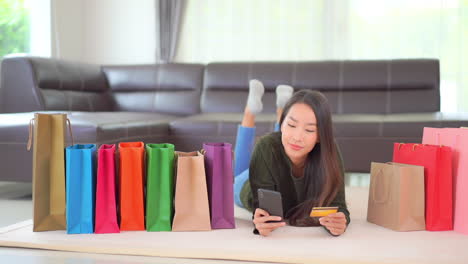 a pretty young woman lying on the floor surrounded by colorful shopping bags double-checks her credit card number with her smartphone screen