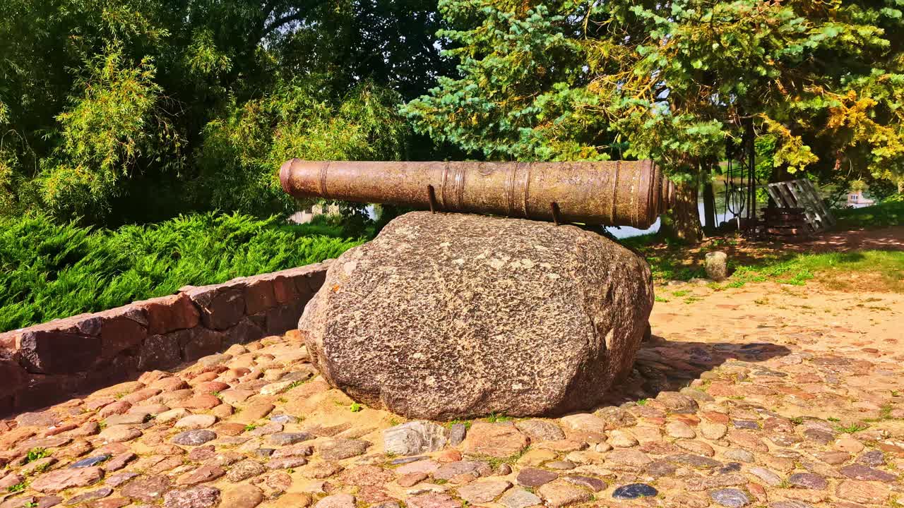A smooth push-in shot focuses on a historic cannon resting on a massive boulder, a monument at the beautiful medieval Jaunpils Castle, a popular tourist destination in the Latvian countryside