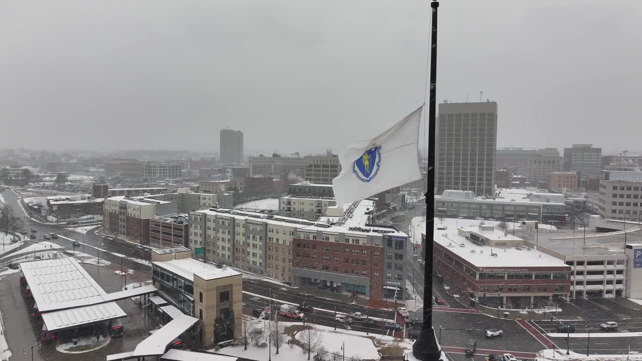 Massachusetts state flag flying over downtown Worcester, MA during blizzard snow storm. Aerial reveal.