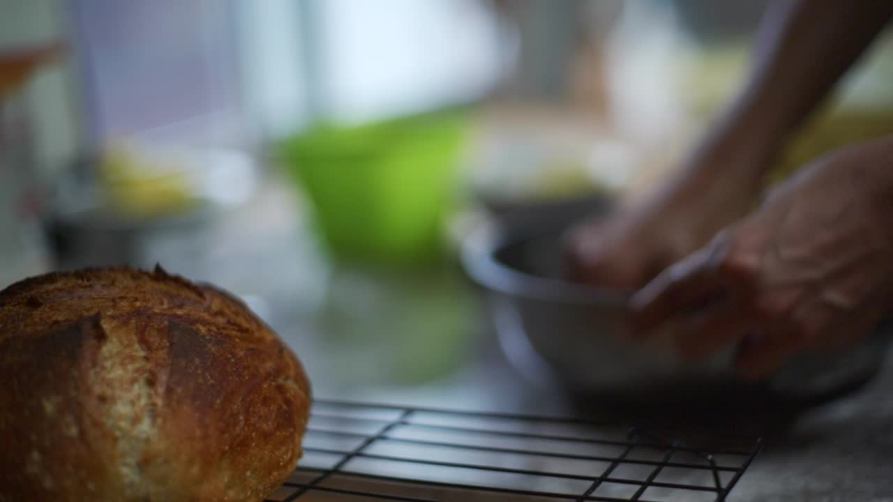 Focus rack from cooked bread roll to uncooked dough being kneaded by hand in steel bowl, filmed as close up slow motion shot