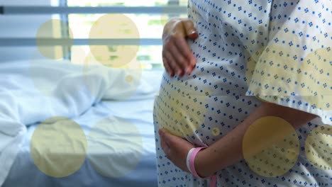 Animation-of-green-spots-over-biracial-pregnant-woman-touching-her-stomach-in-hospital