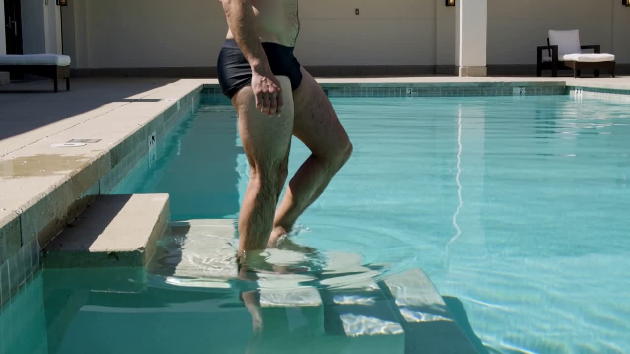 Placing foot onto submerged step, shirtless male in dark briefs descending steps to enter home pool