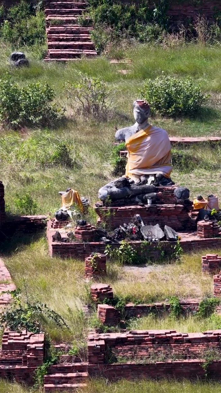 Vertikale Luftaufnahme, die von einer sitzenden Buddha-Statue zu verwitterten Ziegel-Stupas in Thailand schwenkt