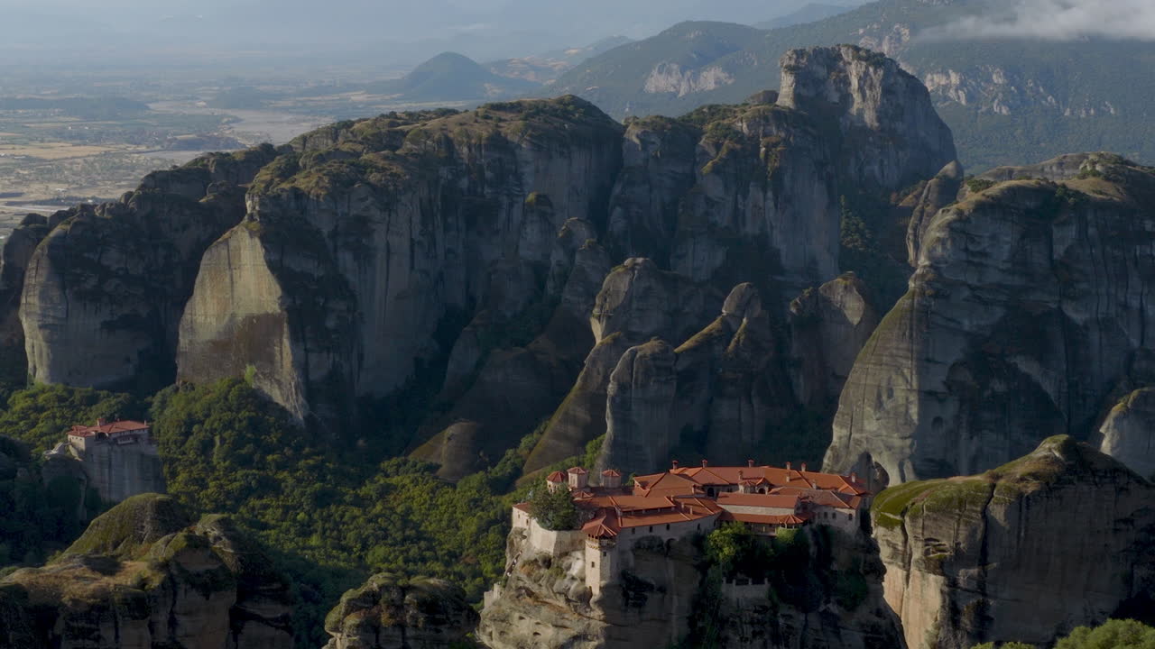 Cinematic aerial view of Meteora monastery in Greece perched on towering cliffs, dramatic rock formations and lush green valley create a breathtaking historic scene