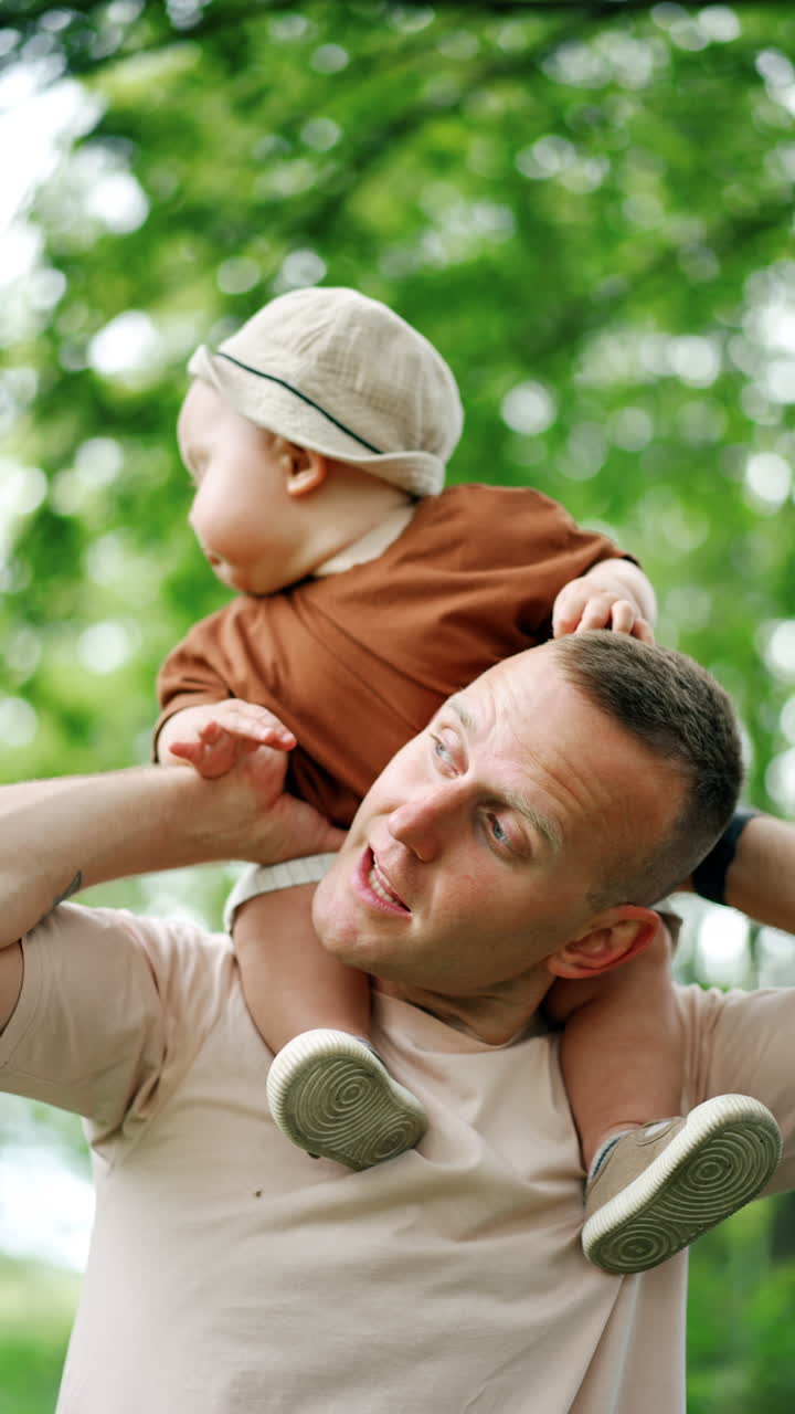 Cute plump baby boy sitting on dad's shoulders tapping him by the head and holding by the nose. Father and son on the walk. Low angle view. Vertical video.