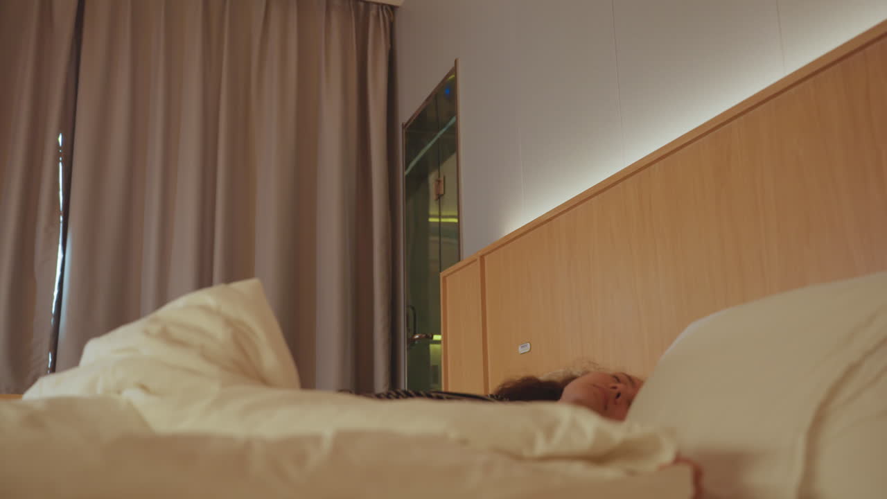 En kvinde sidder og strækker sig i en hotelseng om morgenen, med en varm sengelampe, tunge gardiner trukket for, og krøllet hvidt sengetøj. En fredelig pause med sagte vejrtrækning under et soloophold med en kontemplativ stemning.