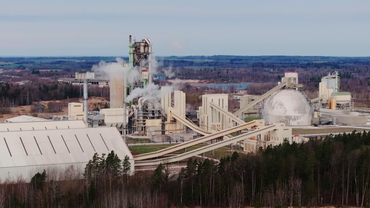 Drone slides left showing gray cement plant conveyors smoke and forest