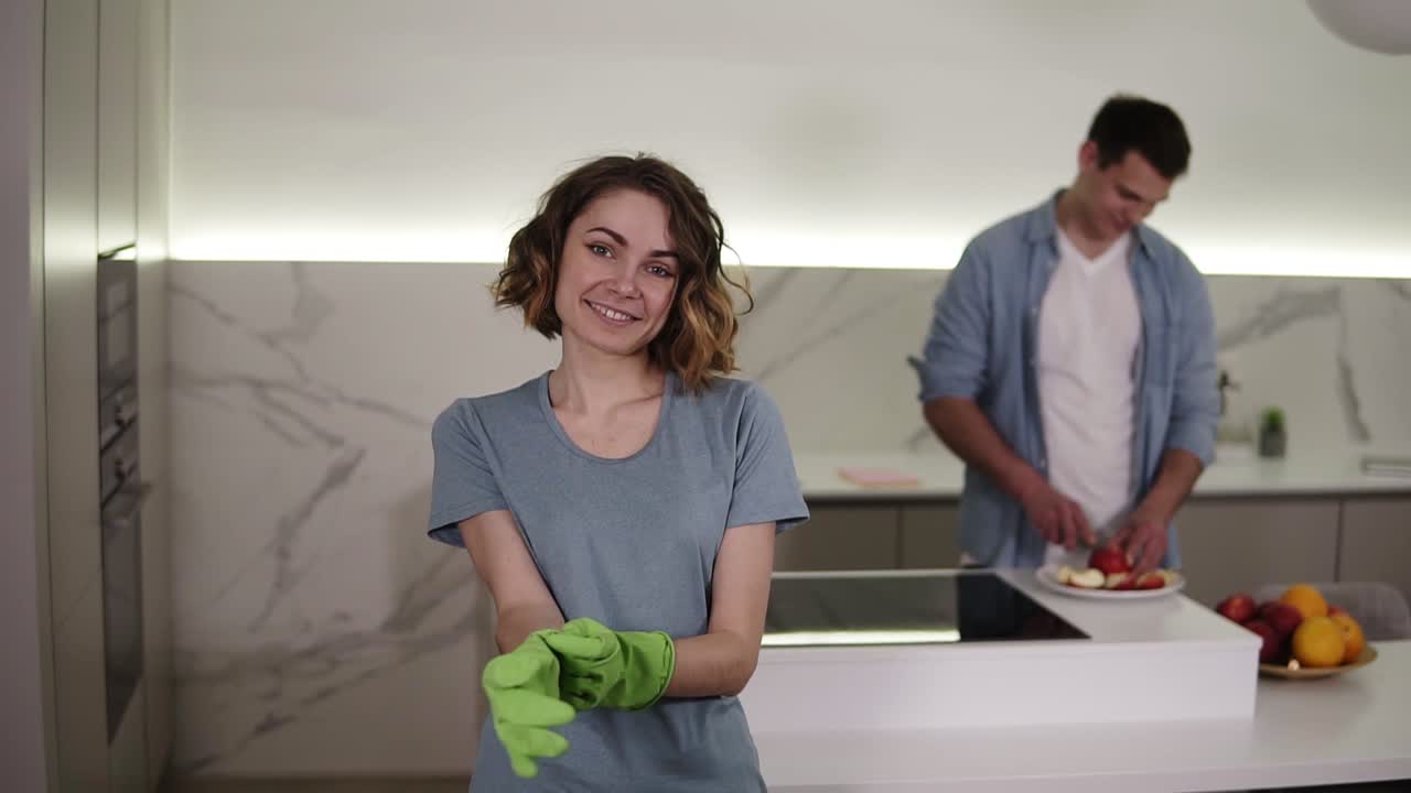Portrait of determined wife ready to start cleaning the household, puts on green gloves and smiling to the camera. On background husband is cutting fruit on a counter on bright, modern kitchen