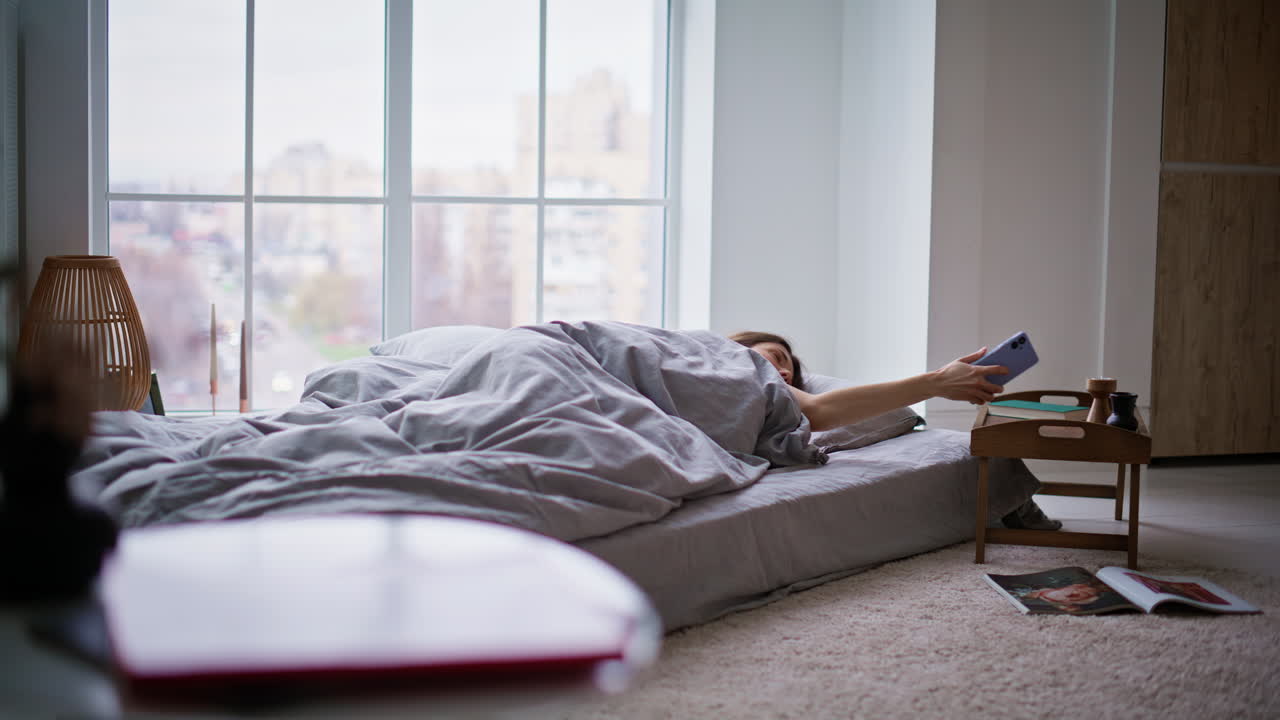 Dovne kvinde sover på madras og tjekker tid på mobiltelefon. Brunette vågner