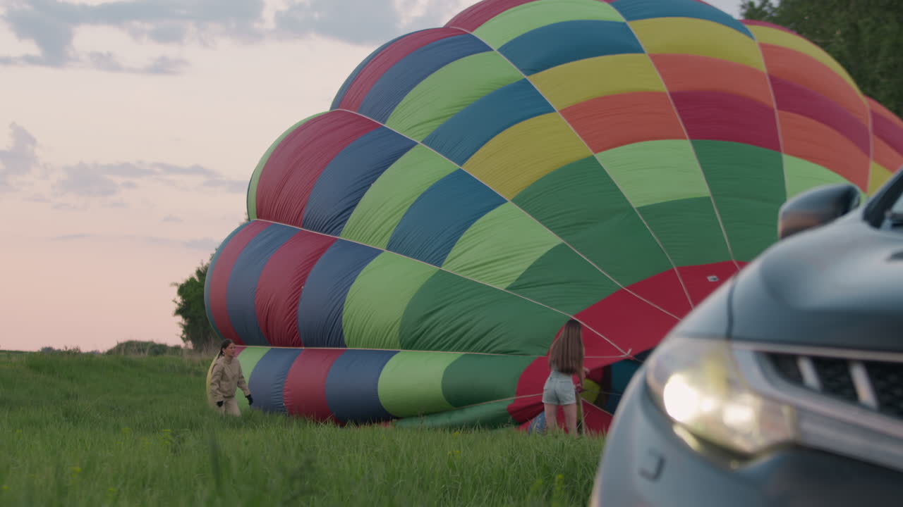 Close up car bumper headlight in foreground framing woman holding hot air balloon envelope panels while another woman runs across tall grass field toward balloon under soft glowing pastel evening sky