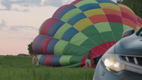 close up car bumper headlight in foreground framing woman holding hot air balloon envelope panels while another woman runs across tall grass field toward balloon under soft glowing pastel evening sky