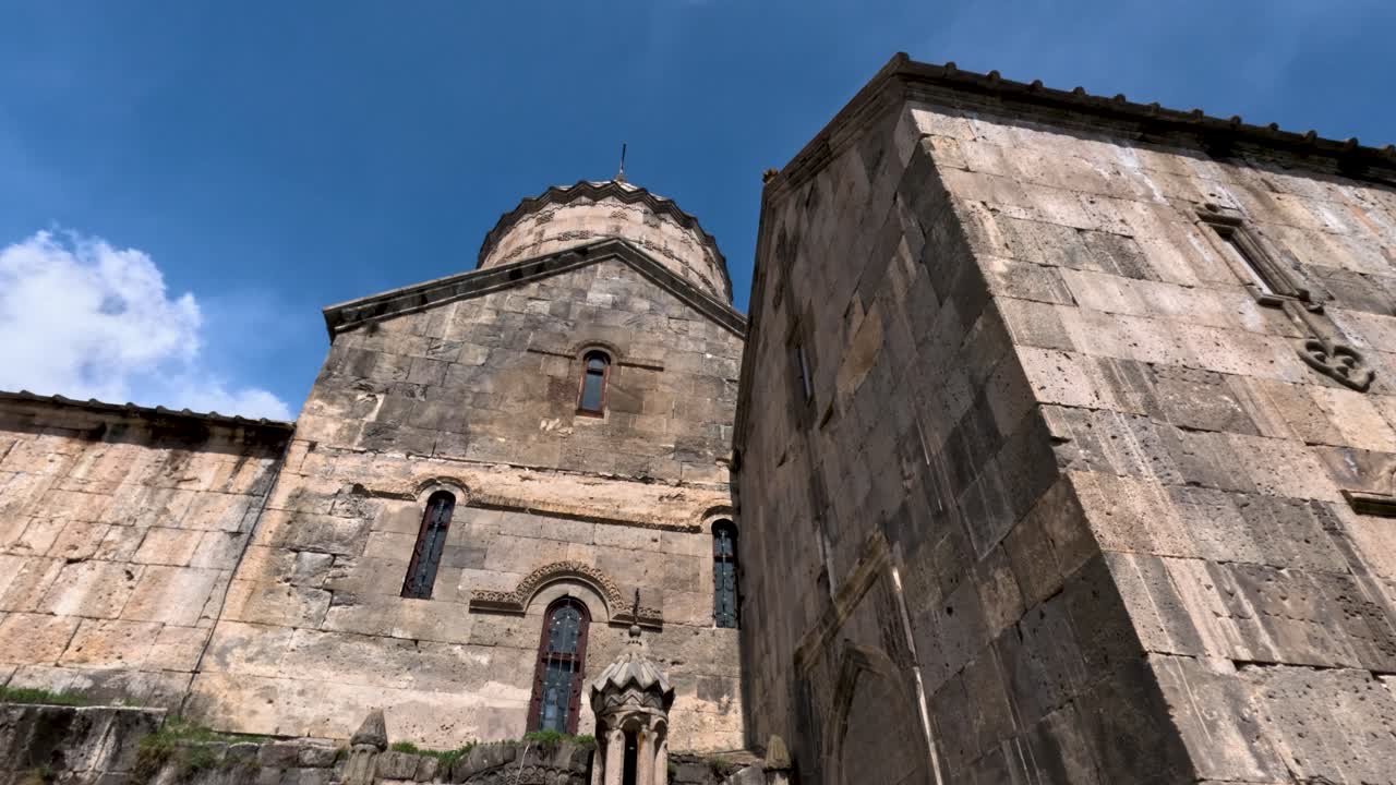 Un visitatore guarda in alto una grande chiesa in pietra in Armenia. Il sole splende luminoso sopra, evidenziando i dettagli della chiesa. L'atmosfera è vivace con turisti intorno.