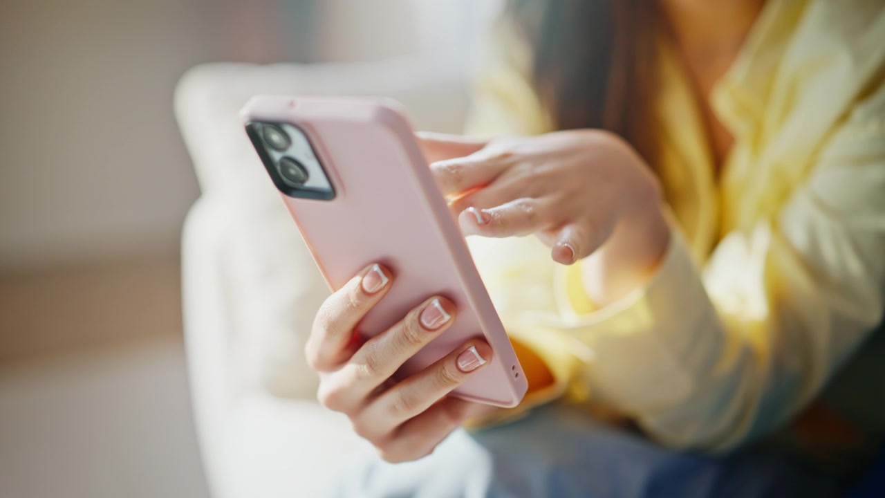 Primo piano delle mani di una donna che tengono uno smartphone rosa. Lei scorre i messaggi a casa